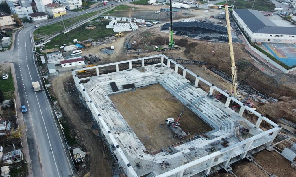 Havadan görünüm: yol kenarında inşaatı süren beton tribünlü saha yapısı ve kule vinç